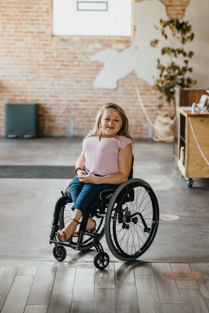 A white woman, Emily, with blonde, shoulder-length hair sits in a black wheelchair. She is short of stature. She is wearing a pink short-sleeved shirt and blue jeans with brown wedge heels. The ground is partially cement, partially wood. There is a brick wall behind her.